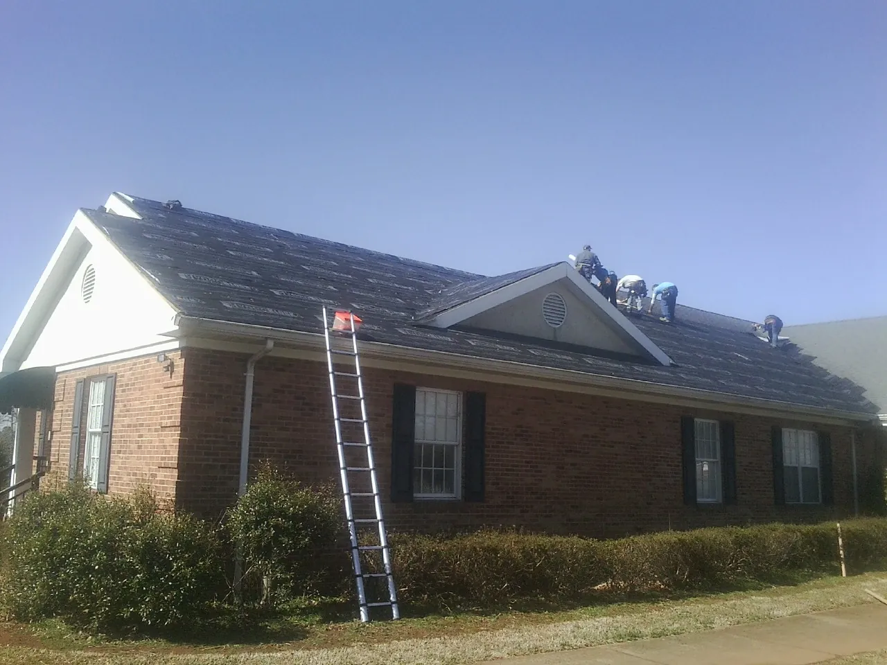 Crew tearing shingles off a brick ranch roof with a ladder set — Emergency Roof Repair work by our crew in Milton